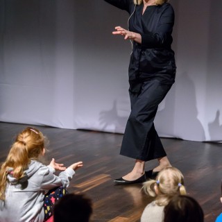 "Pan Dźwięk" - spektakl i warsztaty - Aktorka m&oacute;wi do dzieci siedzących na podłodze i pokazuje gesty rękami.