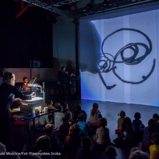 "Pan Dźwięk" - spektakl i warsztaty - Dzieci siedzą na podłodze. Patrzą na ekran, na kt&oacute;rym jest wyświetlany abstrakcyjny obraz.