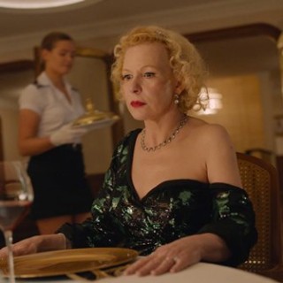 Kadr z filmu W trójkącie - Kobieta w lelganckiej sukni siedzi przy stole. W tle dwie kelnerki.