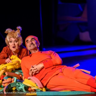 "Wystarczy być" - spektakl dla dzieci Teatru Maska - Kobieta i mężczyzna w przebraniach grają w przedstawieniu dla dzieci na kolorowej scenie. Mężczyzna leży opierając się na łokciu, a kobieta wskazuje na coś palcem. - Fot : Przemysław Sroka