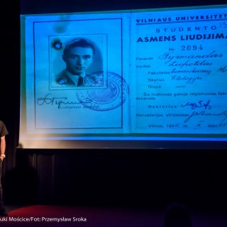 Spotkanie z biografem Leopolda Tyrmanda, Marcelem Woźniakiem - Mężczyzna w okularach mówi do mikrofonu. Na ekranie wyświetla się czarno-białe zdjęcie legitymacji studenckiej.