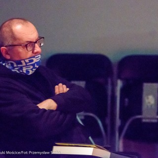 Spotkanie z biografem Leopolda Tyrmanda, Marcelem Woźniakiem - Mężczyzna w chuście i w okularach siedzi na widowni z założonymi rękami.