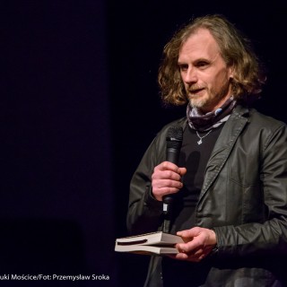 Spotkanie z biografem Leopolda Tyrmanda, Marcelem Woźniakiem - Mężczyzna w czarnej kurtce mówi do mikrofonu trzymając w dłoni książkę.