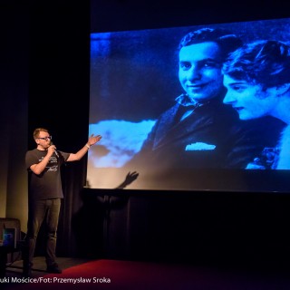 Spotkanie z biografem Leopolda Tyrmanda, Marcelem Woźniakiem - Mężczyzna w okularach mówi do mikrofonu wskazując na ekran. Na ekranie wyświetla się czarno-białe zdjęcie kobiety i mężczyzny.