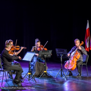 Koncert Patriotyczny Kwartetu Smyczkowego Filharmonii Krakowskiej - Czteroosobowa grupa muzyków gra na instrumentach smyczkowych. W tle widać trzy flagi Polski.