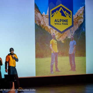 Festiwal Górnolotni 2021. Dzień pierwszy. - Mężczyzna w czapce mówi do mikrofonu. Za nim, na ekranie, wyświetla się zdjęcie dwóch mężczyzn oraz napis: ,,Alpine Wall Tour" - Fot : Przemysław Sroka