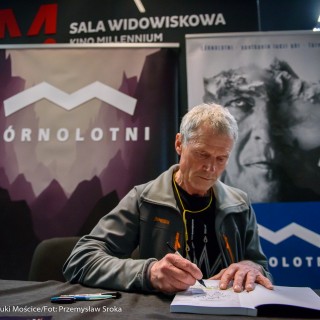 Festiwal Górnolotni 2021. Dzień pierwszy. - Mężczyzna siedzi i podpisuje książkę. Za nim widnieje napis: ,,Górnolotni" - Fot : Przemysław Sroka