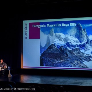 Festiwal Górnolotni 2021. Dzień pierwszy. - Dwóch mężczyzn, w lewym dolnym rogu, siedzi spoglądając na ekran, na którym wyświetla się zdjęcie krajobrazu górskiego oraz napis: ,,Patagonia: Masyw Fitz Roya 1987" - Fot : Przemysław Sroka