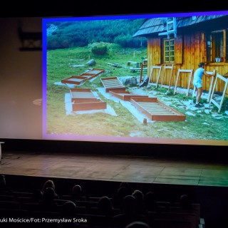 Festiwal Górnolotni 2021. Dzień drugi. - Mężczyzna mówi do mikrofonu i wskazuje na ekran, na którym wyświetla sie zdjęcie drewnianego domu. - Fot : Przemysław Sroka