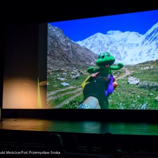 Festiwal Górnolotni 2021. Dzień drugi. - Mężczyzna z mikrofonem siedzi obok ekranu, na którym wyświetla się zdjęcie krajobrazu górskiego i pluszowa żaba. - Fot : Przemysław Sroka