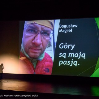 Festiwal Górnolotni 2021. Dzień drugi. - Mężczyzna z mikrofonem stoi obok ekranu, na którym wyświetla się zdjęcie mężczyzny, oraz napis: ,,Bogusław Magrel - Góry są moją pasją" - Fot : Przemysław Sroka