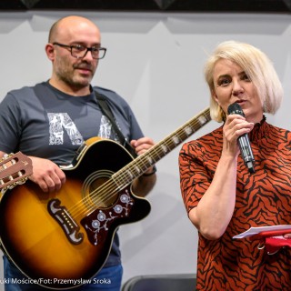 Akcja muzyczna na 50-lecie Centrum - Drektor CSM w pomarańczowej sukience mówi do mikrofonu, za nią stoi mężczyzna z gitarą w rękach.  - Fot. : Przemysław Sroka