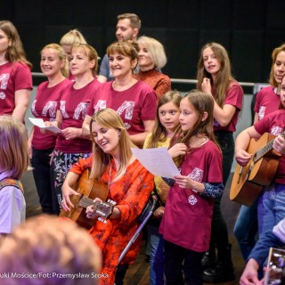 Akcja muzyczna na 50-lecie Centrum - Grupa ludzi w bordowych koszulkach śpiewa, niektórzy trzymają kartki w rękach, przed nimi kobieta w pomarańczowej sukience oraz dzieci grają na gitarach.  - Fot. : Przemysław Sroka