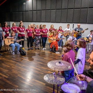 Akcja muzyczna na 50-lecie Centrum - Grupa ludzi stoi w półkole i śpiewa, niektórzy mają bordowe koszulki, dwie kobiety siedzą na krzesłach z gitarami w rękach, na przeciwko grają muzycy.  - Fot. : Przemysław Sroka
