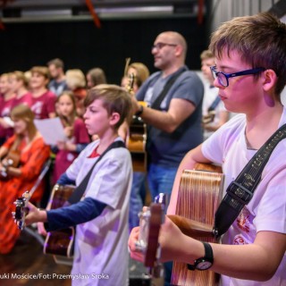 Akcja muzyczna na 50-lecie Centrum - Mężczyzna w okularach oraz dwóch chłopczyków grają na gitarach, za nimi kobieta w pomarańczowej sukience siedzi na krześle z gitarą w ręce, w tle grupa ludzi w bordowych koszulkach trzyma kartki w rękach.  - Fot. : Przemysław Sroka