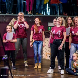 Akcja muzyczna na 50-lecie Centrum - Grupa kobiet w bordowych koszulkach oraz dwójka dzieci stoi na scenie i śpiewa, niektóre z nich mają kartki w rękach, jedna telefon.  - Fot. : Przemysław Sroka
