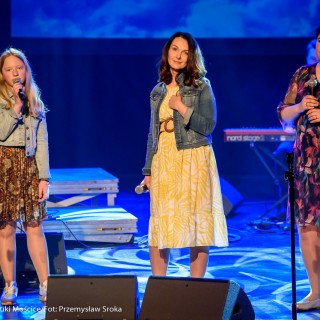 "Znów wędrujemy" - widowisko muzyczne - Dwie kobiety w kwiecistych sukienkach stoją z mikrofonami w ręce, obok ich dziewczynka w kwiecistej sukience śpiewa do mikrofonu.  - Fot. : Przemysław Sroka
