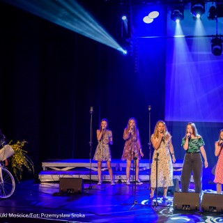 "Znów wędrujemy" - widowisko muzyczne - Grupa kobiet w kwiecistych sukienkach śpiewa do mikrofonów, z boku sceny kobieta stoi przy rowerze w którego koszyku jest bukiet żółtych kwiatów.  - Fot. : Przemysław Sroka
