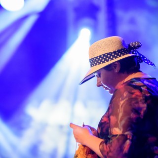 "Znów wędrujemy" - widowisko muzyczne - Kobieta w kwiecistej sukience z kapeluszem na głowie, siedzi z profilu i dzierga coś kolorowego.  - Fot. : Przemysław Sroka