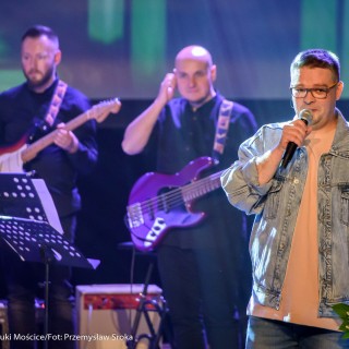 "Znów wędrujemy" - widowisko muzyczne - Mężczyzna w okularach śpiewa, trzyma kwiaty w rękach, za nim dwóch mężczyzn gra na gitarach.  - Fot. : Przemysław Sroka