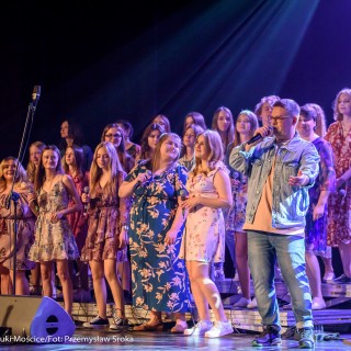 "Znów wędrujemy" - widowisko muzyczne - Grupa kobiet w kwiecistych sukienkach stoi w trzech rzędach na stopniach i patrzy na mężczyznę w okularach, który stoi obok i śpiewa do mikrofonu.  - Fot. : Przemysław Sroka