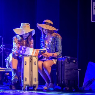 "Znów wędrujemy" - widowisko muzyczne - Dwie kobiety w kwiecistych sukienkach i z kapeluszami na głowach siedzą na ławce, patrzą na rozłożoną mapę Tarnowa, za nimi stoi znak przystanku autobusowego. - Fot. : Przemysław Sroka
