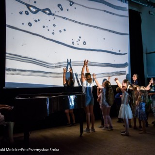 "Pan Dźwięk" - spektakl i warsztaty - Grupa dzieci na scenie zwrócone do ekranu rzutnika na którym wyświelają się wzory, niektóre z dzieci unoszą ręce, kobieta w jasno-różowej sukience siedzi przy fortepianie.  - Fot. : Przemysław Sroka