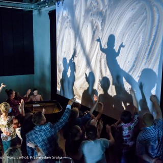 "Pan Dźwięk" - spektakl i warsztaty - Grupa dzieci i dorosłych na scenie stoją przed ekranem rzutnika, unoszą ręce i tworzą na nim cienie, kobieta w jasno-różowej sukience siedzi przy fortepianie.  - Fot. : Przemysław Sroka