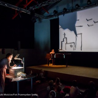 "Pan Dźwięk" - spektakl i warsztaty - Kobieta rysuje na ekranie urządzenia postawionego na stoliku, przed nią na poduszkach na podłodze siedzą dzieci i patrzą na ekran rzutnika wywieszonego na scenie na którym pojawiają się rysowane przez kobietę obrazki, druga kobieta siedzi przy fortepianie na scenie. - Fot. : Przemysław Sroka