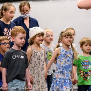 "Pan Dźwięk" - spektakl i warsztaty - Grupa dzieci stoi w dwóch rzędach, trzymając się za ręce i patrzy na kobietę w jasno-różowej sukience, za nimi stoi dwójka dorosłych, jedna kobieta ma maseczkę.  - Fot. : Przemysław Sroka