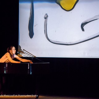 "Pan Dźwięk" - spektakl i warsztaty - Kobieta na scenie nachyla się do strun fortepianu, spogląda do góry na ekran rzutnika na którym widać namalowane kreski.  - Fot. : Przemysław Sroka