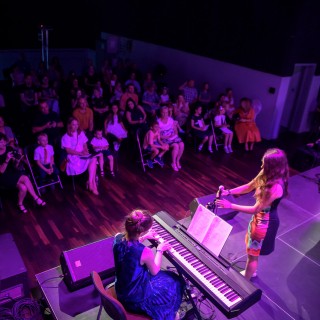 MiniLab - zakończenie roku 2020/2021 - Ludzie na widowni oglądają dwie kobiety na scenie; jedna z nich trzyma mikrofon, druga gra na keyboardzie.  - Fot. :Przemysław Sroka