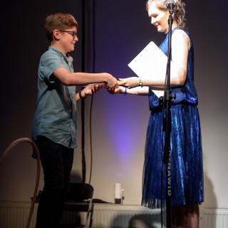 MiniLab - zakończenie roku 2020/2021 - Kobieta w niebieskiej sukience stoi na scenie, w jednej ręce trzyma dyplom, a drugą ściska rękę chłopczyka w koszuli i okularach, oboje uśmiechają się. - Fot. :Przemysław Sroka