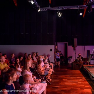 MiniLab - zakończenie roku 2020/2021 - Ludzie na widowni patrzą na kobietę w czerwonej sukience, która stoi na scenie i mówi do mikrofonu.  - Fot. :Przemysław Sroka