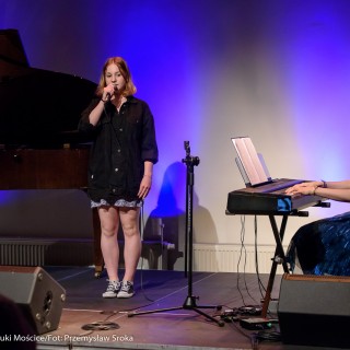MiniLab - zakończenie roku 2020/2021 - Kobieta w sukience i czarnej katanie śpiewa do mikrofonu na scenie, a kobieta w niebieskiej sukience gra na keyboardzie.  - Fot. :Przemysław Sroka