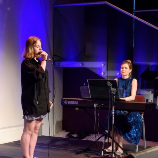 MiniLab - zakończenie roku 2020/2021 - Kobieta w sukience i czarnej katanie śpiewa do mikrofonu na scenie, a kobieta w niebieskiej sukience gra na keyboardzie.  - Fot. :Przemysław Sroka