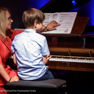 MiniLab - zakończenie roku 2020/2021 - Kobieta w czerwonej sukience pokazuje nuty chłopczykowi, który siedzi przy fortepianie.  - Fot. :Przemysław Sroka