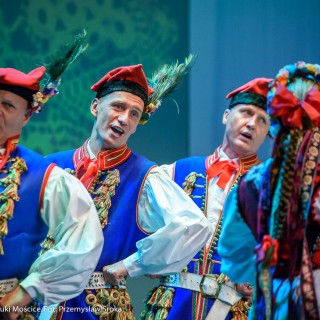 Świerczkowiacy - zakończenie sezonu artystycznego 2020/2021 - Grupa mężczyzn w niebieskich strojach ludowych z czapkami z piórem na głowach, tańczą i śpiewają na scenie. - Fot. :Przemysław Sroka