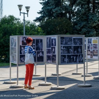 Mościce mam w sercu - wystawa - Kobieta w czerwonych włosach ogląda wystawę zdjęć ustawioną na zewnątrz. - Fot. :Przemysław Sroka