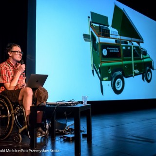 Festiwal Dalekosiężni 2021. Dzień pierwszy. - Mężczyzna na wózku inwalidzkim z laptopem mówi do mikrofonu. Za nim znajduje się ekran ze zdjęciem przedstawiającym pojazd terenowy. - Fot. :Przemysław Sroka
