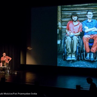 Festiwal Dalekosiężni 2021. Dzień pierwszy. - Mężczyzna na wózku inwalidzkim z laptopem mówi do mikrofonu. Obok niego znajduje się ekran ze zdjęciem przedstawiającym dwoje mężczyzn na wózkach inwalidzkich. - Fot. :Przemysław Sroka