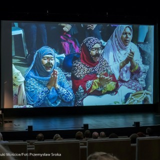 Festiwal Dalekosiężni 2021. Dzień pierwszy. - W lewym dolnym rogu kobieta mówi do mikrofonu. Na ekranie pokazane jest zdjęcie trzech kobiet ubranych w ludowe stroje, składających ręce do modlitwy. - Fot. :Przemysław Sroka