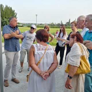 Historie z Mościc w opowieściach lokalnych przewodników - Grupa ludzi stoi przed wystawą zdjęć i słuchają mężczyzny w kraciastej koszuli, który mówi do mikrofonu. - Fot. Dorota Omylska-Bielat