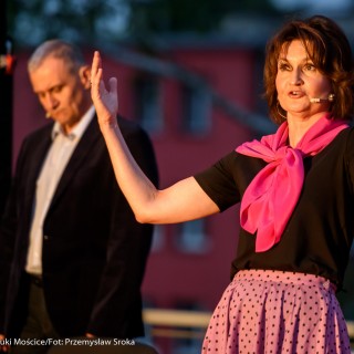 "Małe zbrodnie małżeńskie" - Muzyczne Tarasy 2021 - Kobieta w czarnej bluzce i różowej chustce mówi do mikrofonu z uniesionymi rękami, za nią stoi mężczyzna w marynarce. - Fot.: Przemysław Sroka