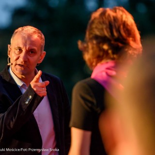 "Małe zbrodnie małżeńskie" - Muzyczne Tarasy 2021 - Mężczyzna w marynarce mówi do mikrofonu i pokazuje palcem wskazującym na kobietę w czarnej bluzce i różowej chustce, która stoi przed nim. - Fot.: Przemysław Sroka