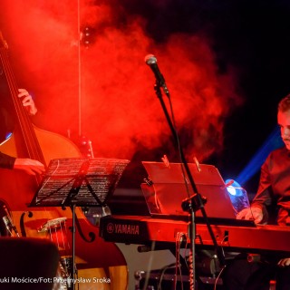 "Małe zbrodnie małżeńskie" - Muzyczne Tarasy 2021 - Mężczyźni siedzą na krzesłach, jeden gra na keyboardzie, drugi na wiolonczeli, nad nimi unosi się czerwony dym. - Fot.: Przemysław Sroka