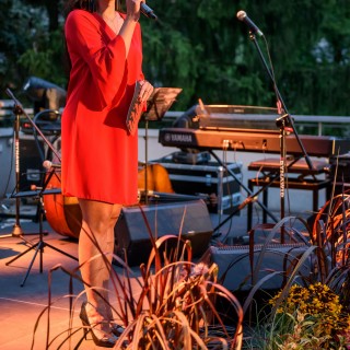 "Małe zbrodnie małżeńskie" - Muzyczne Tarasy 2021 - Kobieta w czerwonej sukience stoi na scenie i mówi do mikrofonu, przód sceny ozdobiony jest kwiatami. - Fot.: Przemysław Sroka