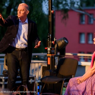 "Małe zbrodnie małżeńskie" - Muzyczne Tarasy 2021 - Kobieta w czarnej bluzce i różowej chustce siedzi na krześle na scenie i patrzy na mężczyznę w marynarce, który mówi do mikrofonu i wskazuje gdzieś ręką. - Fot.: Przemysław Sroka