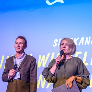Festiwal Tarnowianie - dzień 1 - Kobieta i mężczyzna na scenie, kobieta mówi do mikrofonu, za nimi wyświetlają się napisy na dużym ekranie.  - Fot. Przemysław Sroka