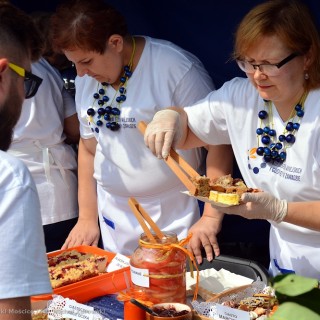 Festiwal Tarnowianie - dzień 2 - Na stoisku grupa kobiet w białych bluzkach i niebieskich koralach, jedna z nich nakłada ciasta na tackę mężczyźnie, który stoi po drugiej stronie lady.  - Fot. Przemysław Sroka i Michał Żurowski